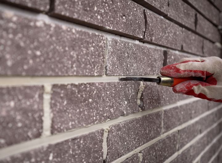 A gloved hand raking out the mortar joint between dark bricks with a pointing tool.